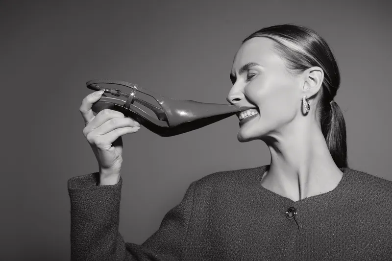 Smiling white woman with fair skin holds high heel shoe near face, playful expression, studio portrait in black and white, fashion and beauty concept.