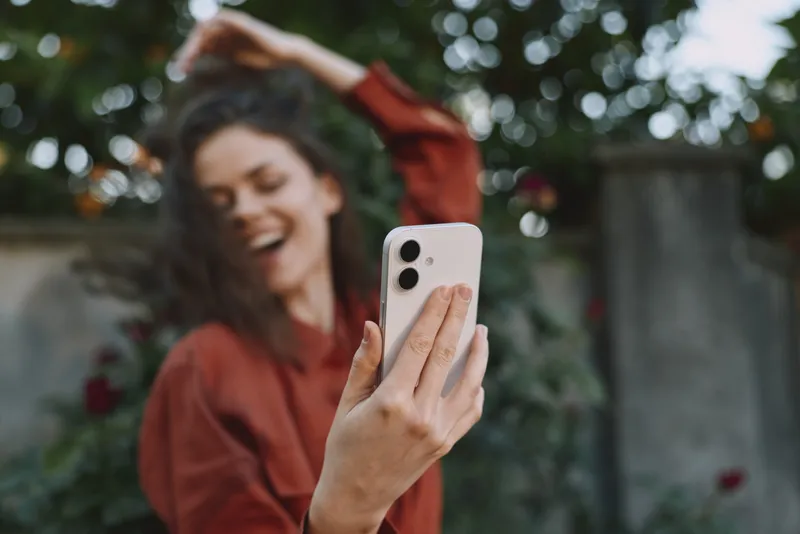 Smiling woman outdoors holds a selfie with a phone, casual attire, bright natural light, blurred garden background, lifestyle portrait capturing joy and candid moment