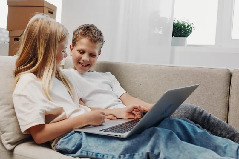 Two children sitting on a cozy sofa smiling while looking at a laptop screen together in a bright living room. Concept of friendship, technology, and learning at home.