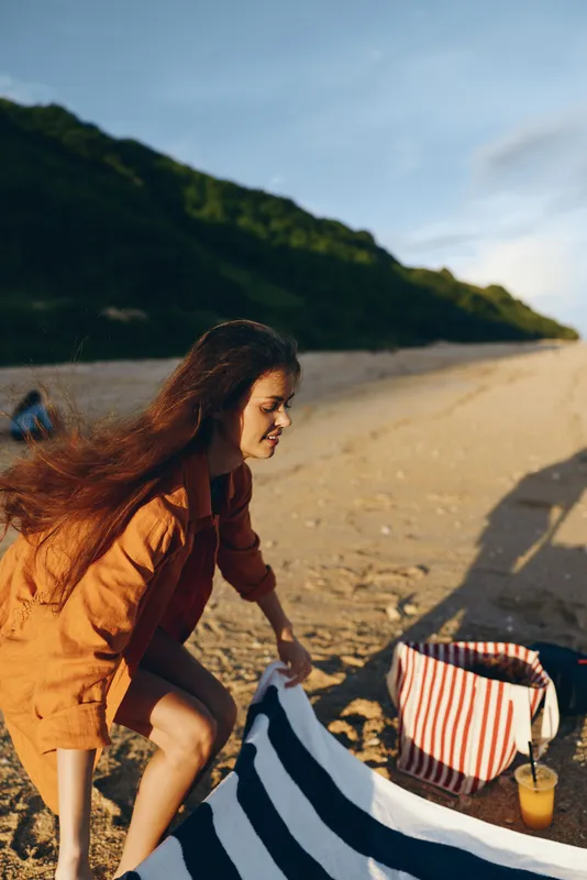 Woman, beach, outdoors, sunlight, windy, walking, striped bag, drink, sand, tote and towel nearby, warm jacket, relaxed posture, mountain coastline background, natural colors