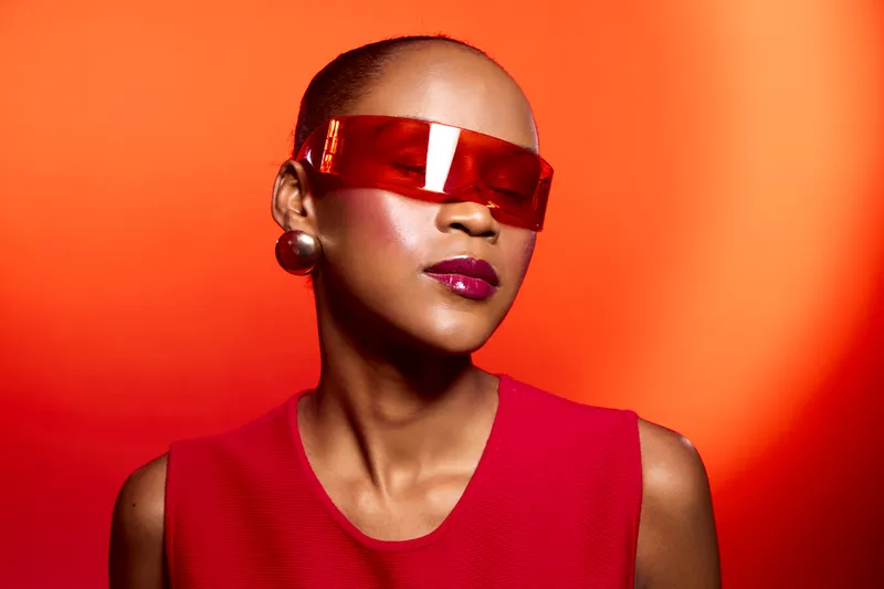 Portrait of a confident black woman with dark skin wearing red futuristic sunglasses and a red sleeveless top against a vibrant red background. Concept of fashion and modern style.