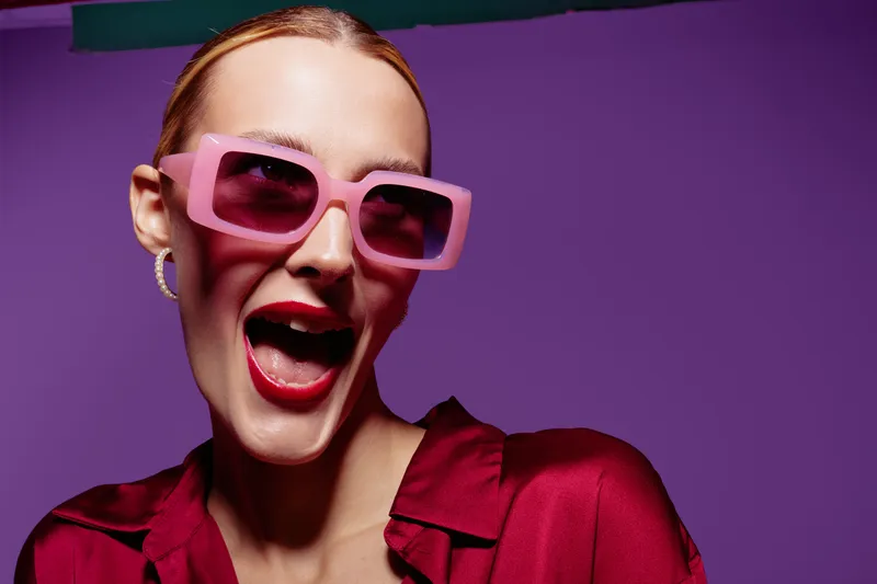 Happy woman with pink sunglasses and red lipstick smiles widely against purple background. Fashion concept with vibrant colors and joyful expression in studio lighting.