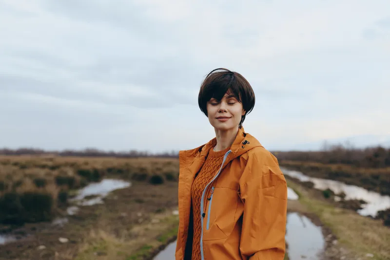 Woman lifestyle outdoors, portrait by a river, orange jacket, autumn nature landscape, cloudy daylight, gentle smile, relaxed casual pose, scenic countryside, bright soft natural colors