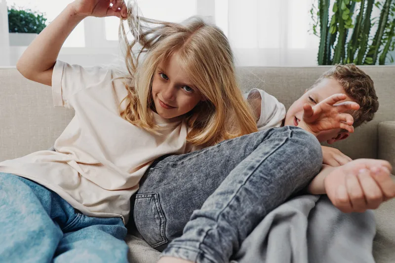 Two playful children sitting on a couch in a bright living room, one girl teasing her brother by pulling his hair. Concept of sibling interaction, family fun, and childhood moments.