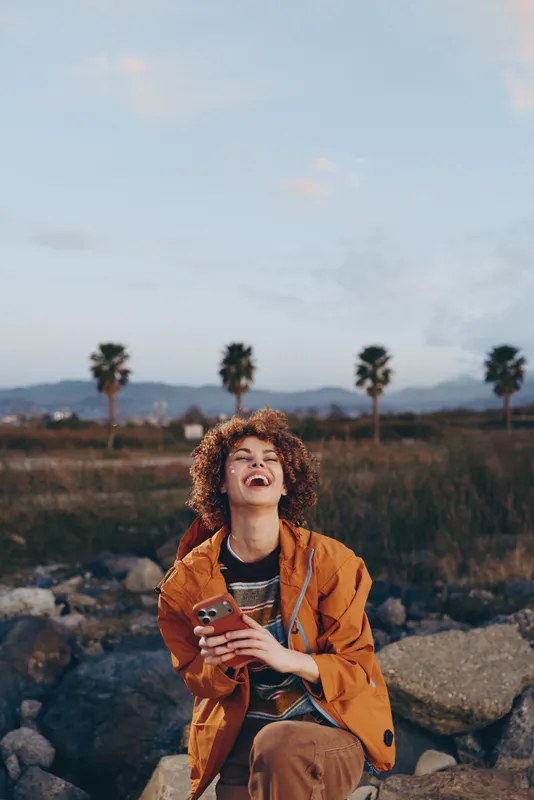 Woman lifestyle portrait outdoors with curly hair, smiling in orange jacket, sitting on rocks and holding phone, palm trees and landscape behind, calm evening mood.