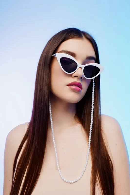 Young woman with long hair wearing white cat eye sunglasses and pearl chain poses against blue background. Concept of beauty, fashion, style, and modern accessories.