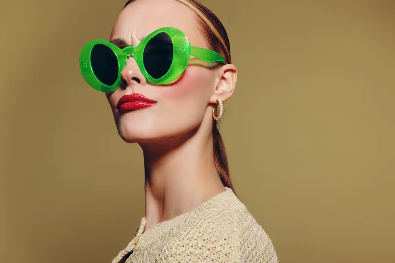 Stylish woman wearing large green sunglasses and red lipstick poses confidently against beige background. Fashion portrait with trendy accessories and bold makeup look.