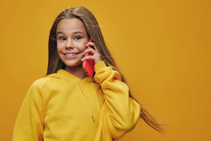 Girl in yellow hoodie talking on red smartphone with smile looking at camera, casual style with long hair on yellow background for communication or technology concept.