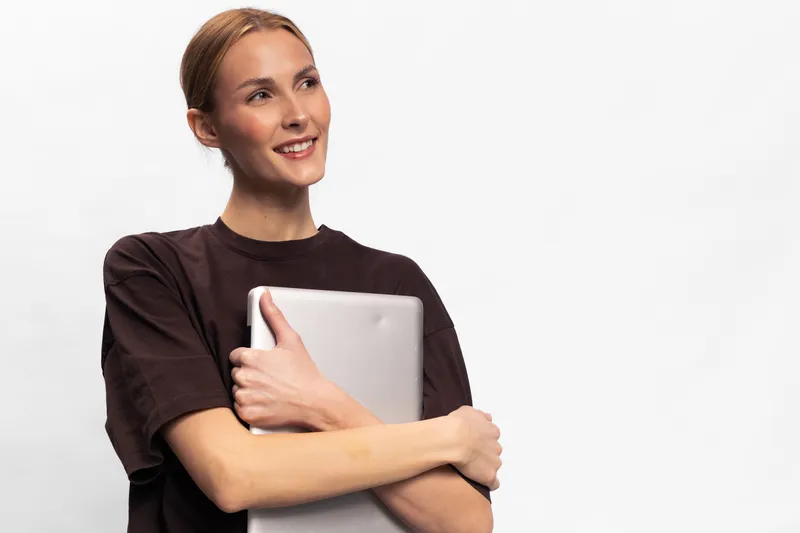 Smiling young woman in casual brown shirt holding a laptop, looking away. Studio portrait on white background. Concept of technology, education, and positive emotions.