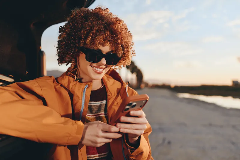 Woman lifestyle, smiling curly hair woman in tan jacket, holding smartphone outdoors near seaside at sunset, warm light, candid travel moment, coastal road, soft focus background, sunlit air