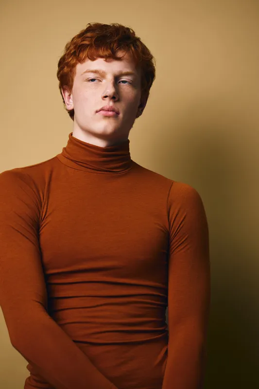 Young man with red hair wearing brown turtleneck sweater posing and looking sideways against yellow background in studio portrait for fashion and style concept.