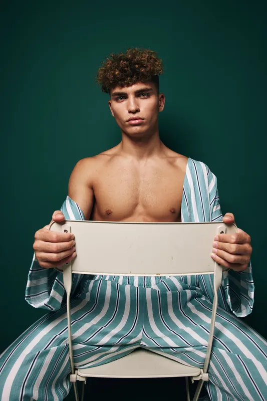 Young man with curly hair wearing striped pajamas sitting backwards on chair against dark green background with serious expression and bare chest visible.