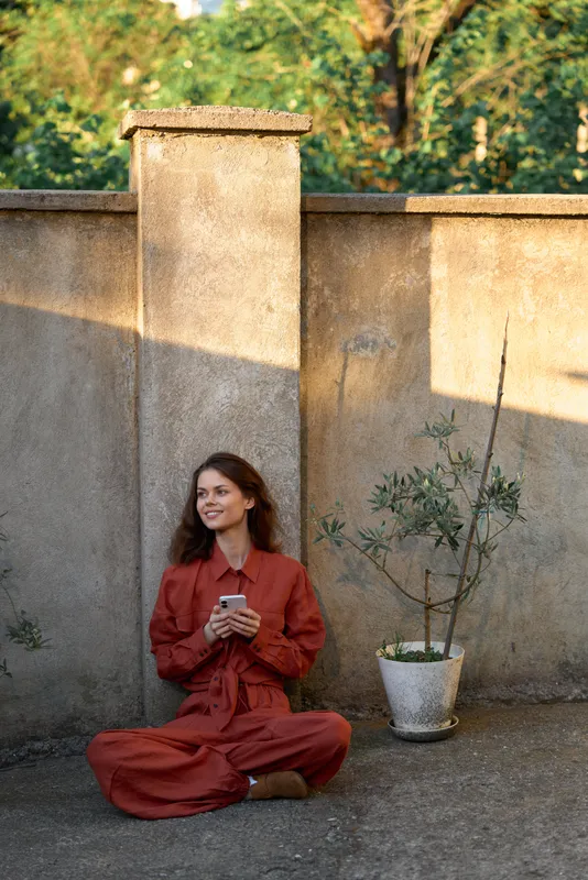 Cheerful woman outdoors, seated cross legged, rust colored outfit, holding a cup, sunlit moment, relaxed pose, natural colors, lush greenery background