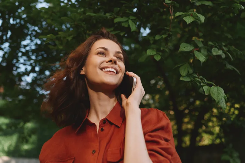 Joyful woman in a red shirt enjoys a sunny outdoor moment among lush trees, smiling warmly in a candid lifestyle portrait set in nature