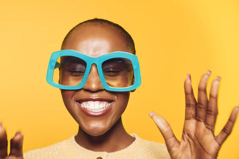 Happy African woman with dark skin wearing oversized blue sunglasses smiles broadly against yellow background. Concept of joy, fashion, and positive lifestyle for dark skinned people.