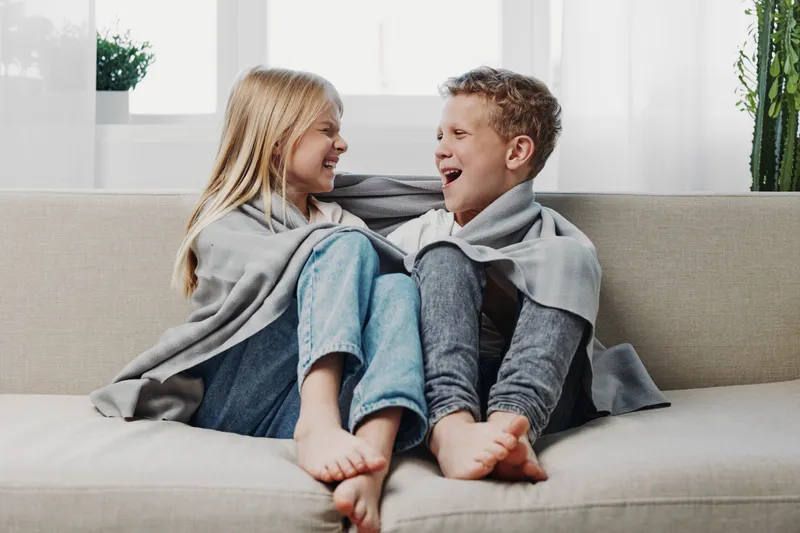 Two children wrapped in a grey blanket sit barefoot on a sofa laughing and looking at each other in a bright cozy living room environment.