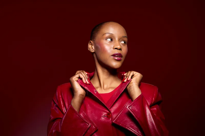 Confident dark skinned African woman wearing red leather jacket looks sideways with thoughtful expression. Studio portrait on dark red background. Concept of fashion, beauty, and style.