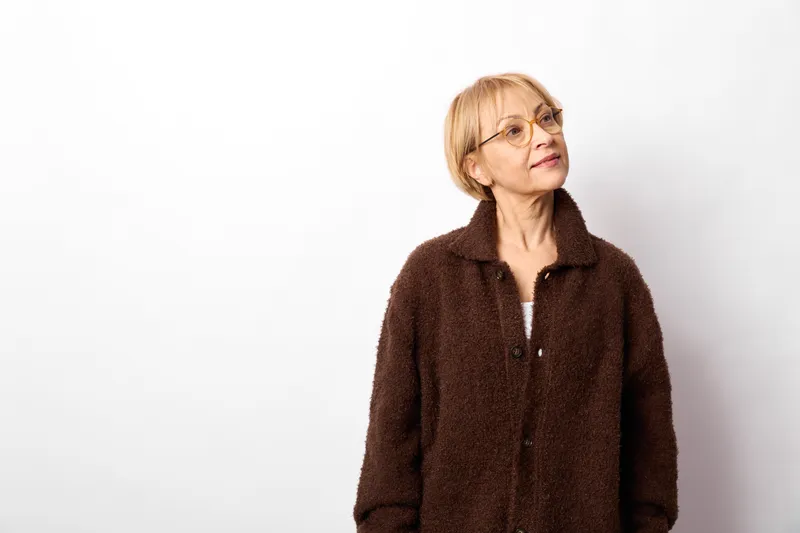 woman senior smiling glasses portrait jacket cozy isolated on white background, short blond hair, dark jacket collar, calm studio head and shoulders