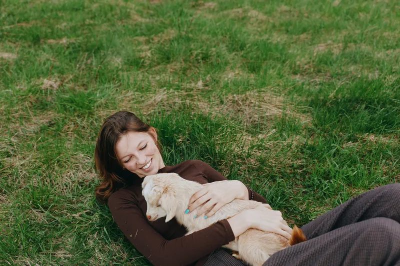 A woman lying on green grass in casualwear, cuddling a small dog and smiling softly. An outdoor moment of relaxation, companionship, and emotional wellbeing, capturing bonds between people and pets