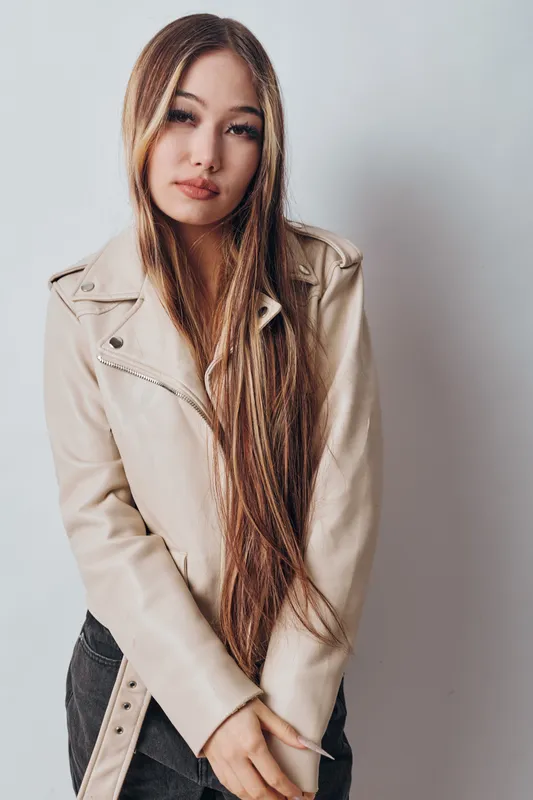 Young woman with long hair wearing a beige jacket and black pants standing against white background. Natural expression, casual fashion, and modern style with light makeup and relaxed pose.