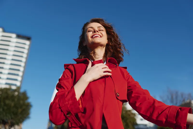 Young woman with curly hair enjoys fresh air outdoors wearing a red coat and smiles with closed eyes under a clear blue sky.