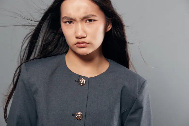 Young woman with long dark hair and serious face wearing buttoned gray jacket on gray background, looking directly at camera with furrowed brows and intense expression.