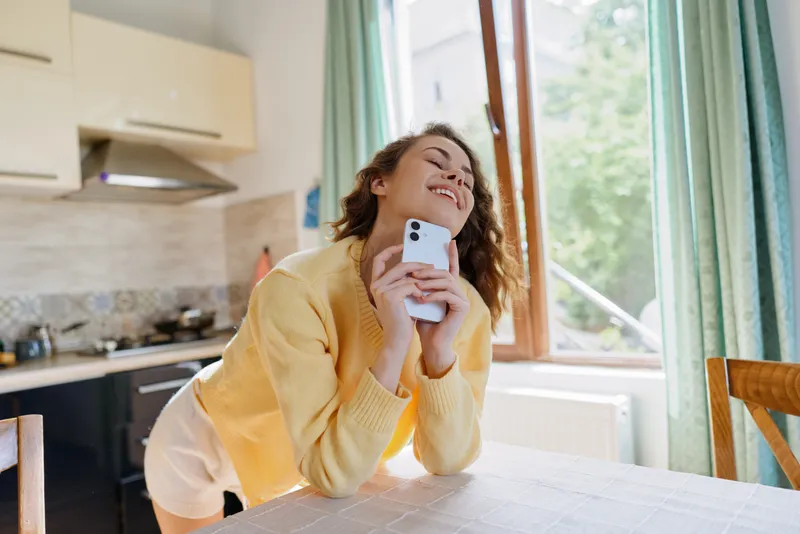 Woman in a cozy kitchen leans on a table, smiling while holding a smartphone, enjoying a relaxed lifestyle moment at home, warm daylight streams through the window