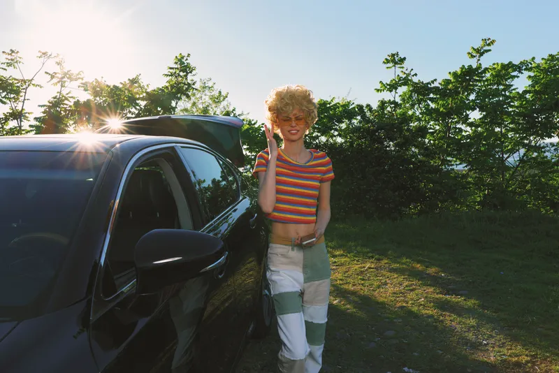 A young blonde woman with curly hair stands beside a dark car in a sunny parking area, flashing a peace sign as she leans against the vehicle in a striped crop top and patchwork pants.