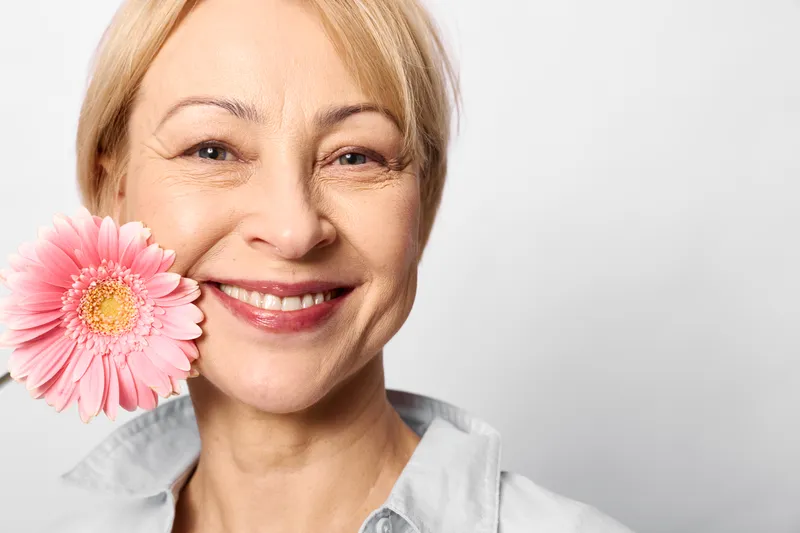 Smiling woman, red hair, holding pink flower near cheek, closeup on white background, skincare wellness selfcare theme, studio soft light, fresh happy mood portrait