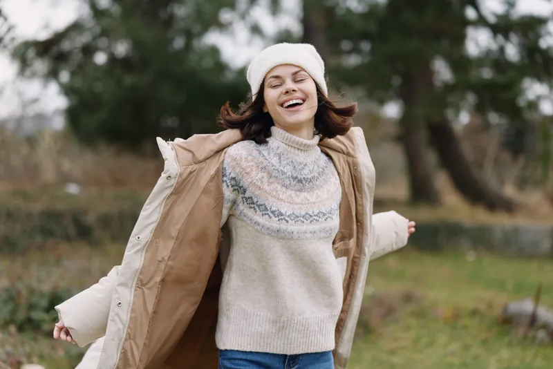 Joyful woman outdoors wearing a warm coat and sweater, smiling as she moves freely in a park breeze, autumn light and candid mood, casual fashion in motion