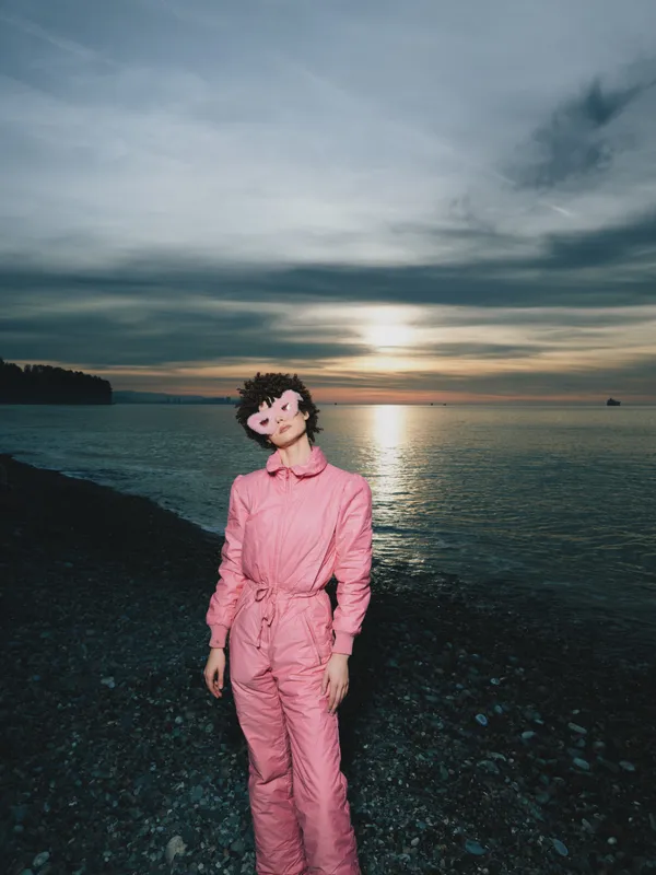 Woman in pink jumpsuit stands on a rocky coastline at sunset, calm sea behind, warm light on her face, relaxed pose, outdoor portrait with dramatic sky and moody colors.