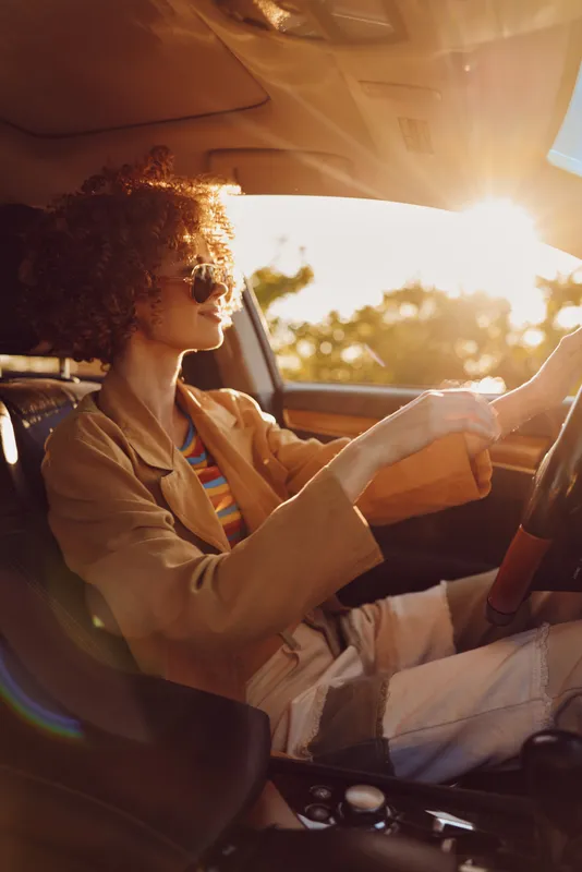 Woman with curly hair driving a car at sunset, warm light on her face, casual outfit, lifestyle moment, relaxed mood, retro film color, sense of freedom.