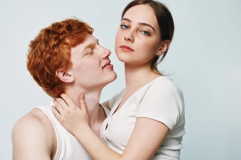 Young couple with red hair and blue eyes in casual white tank tops embracing and looking at camera, indoor portrait with plain light background and natural expression.