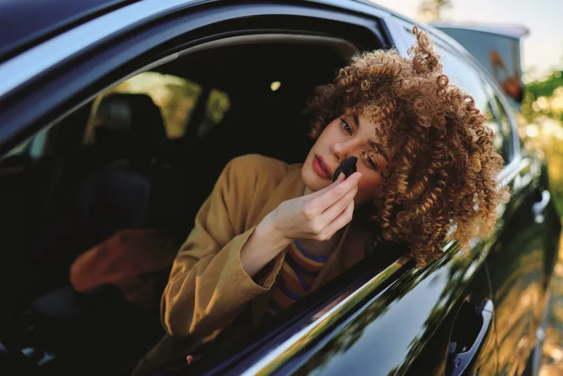 Woman leaning from car window applying makeup, casual lifestyle moment with retro color tones, candid portrait outside vehicle, relaxed mood, warm light and everyday glamour