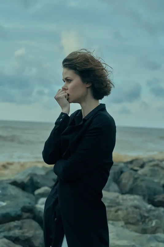 Woman on a rocky coast wearing a dark coat, wind in her hair, a thoughtful pose beside the sea under a moody sky, suitable for fashion or travel visuals
