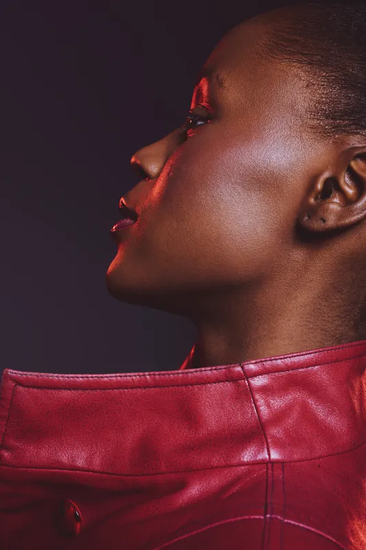 Profile of a dark skinned African woman wearing a red leather jacket. Studio portrait with dramatic lighting highlights smooth skin and elegant features. Concept of fashion and beauty.