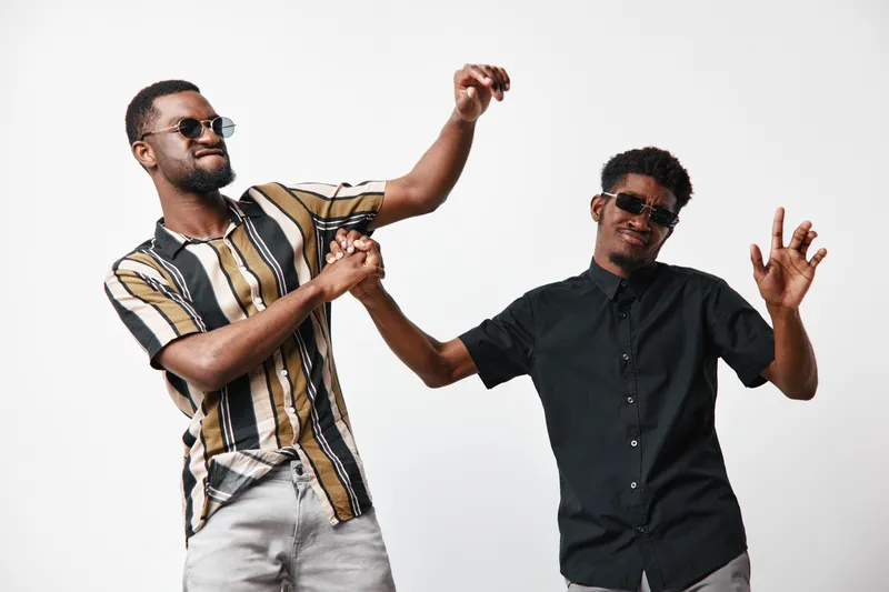 Two young black men with dark skin wearing sunglasses and casual clothes dancing and having fun together on white background. Friendship, joy and celebration with happy emotions.