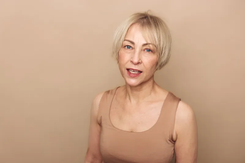 Portrait of smiling mature woman with short blonde hair wearing beige tank top against soft beige background. Concept of natural beauty, aging gracefully, confidence and simplicity.