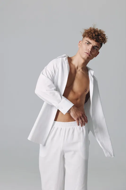 Young man with curly hair wearing white open shirt and pants posing against gray background, casual style and confident expression in studio portrait photography.