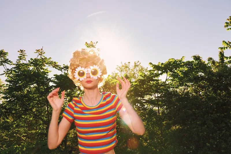 Joyful woman outdoors, wearing a striped top and sunflower sunglasses, basking in warm sunlight, retro color tones, carefree pose, lush greenery background, bright and sunny mood