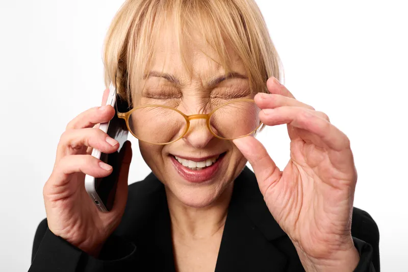 Woman with blonde hair shows stress in a phone call, overwhelmed behind glasses, hands at her ears, eyes squinting, isolated on white office background, hearing anxiety, callcenter communication