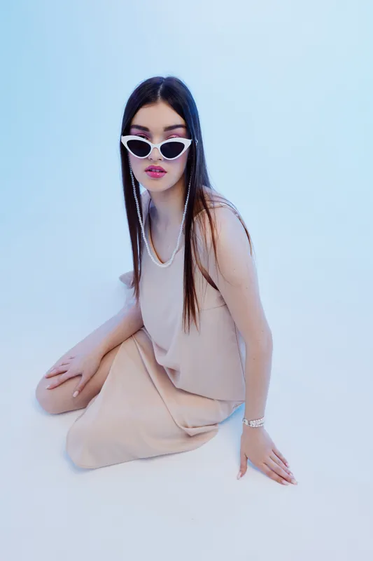 Young woman in beige dress and white sunglasses poses sitting on floor against light blue background. Concept of beauty fashion style and modern elegance with minimalistic studio lighting.