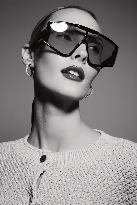 Young white woman with pale skin wears large sunglasses and knitted sweater, posing in studio with confident expression and modern style.