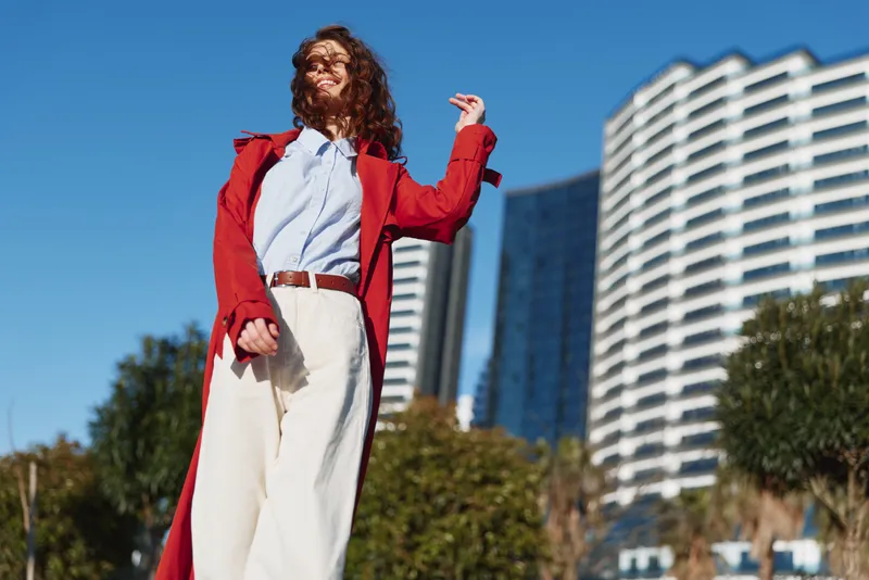 Young woman with curly hair stands outdoors in a red coat and white wide trousers, striking a confident pose against modern buildings on a sunny day.