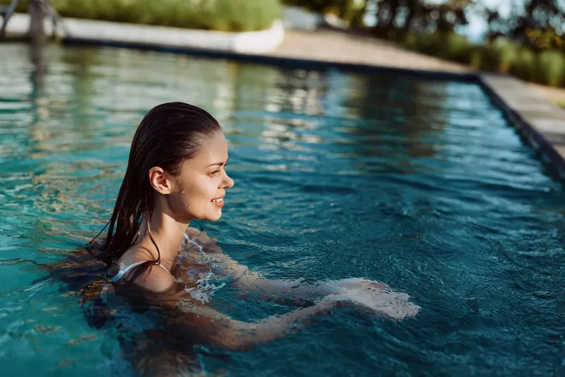 Young woman with wet hair enjoying a swim in an outdoor pool on a sunny day. Relaxed lifestyle concept with natural light and clear blue water creating a peaceful mood.