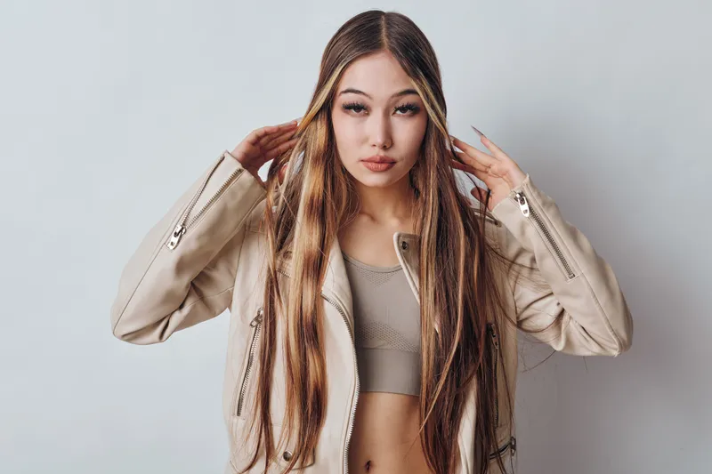 Young woman with long brown hair wearing a beige jacket and crop top posing in front of gray background, fashion style, portrait and confident expression.