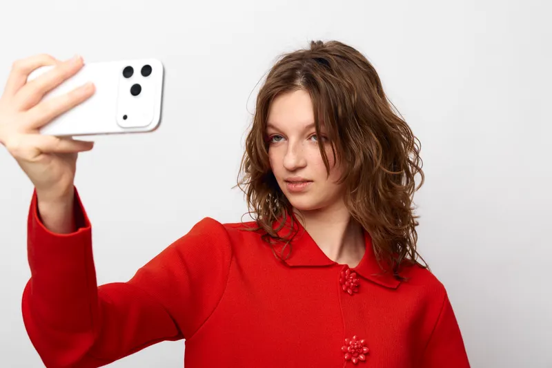 Young woman with curly hair wearing red sweater takes selfie with smartphone on white background. Casual style, natural expression, modern technology and social media concept.