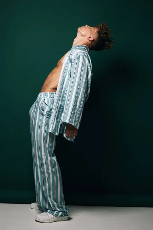 Man in striped pajamas leaning backward with eyes closed against dark background, casual homewear, relaxed posture, curly hair, barefoot on white floor, indoor studio setting.