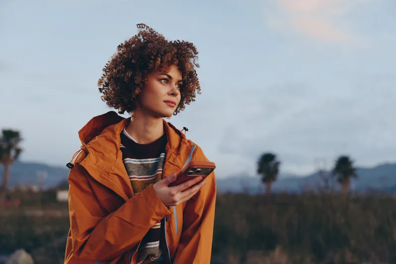 Woman outdoors lifestyle, curly hair and orange jacket, holding a phone while walking, calm confident portrait in warm sunset light, soft breeze, distant trees and hills background