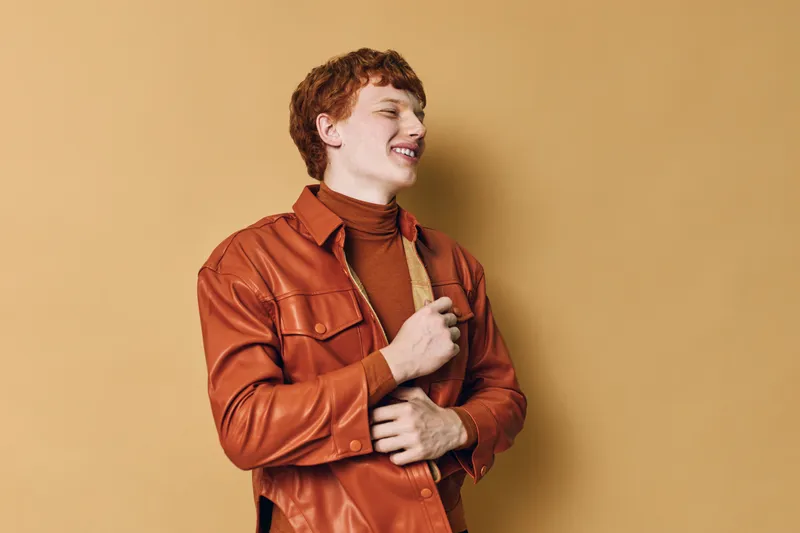 Young man with red hair and leather jacket smiling and looking side on beige background. Casual fashion portrait of male model with relaxed expression and stylish outfit in studio setting.